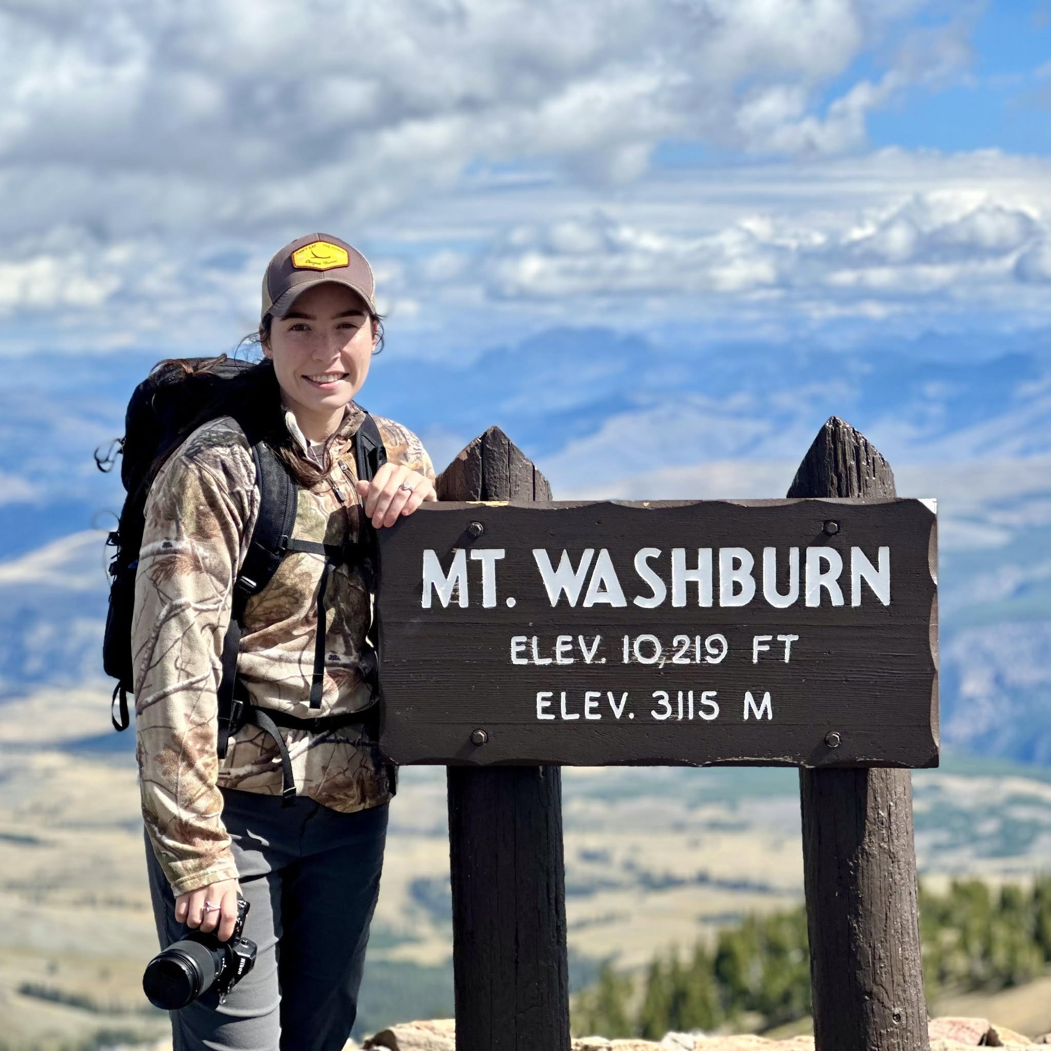 Meredith Lewis at the Mt. Washburn summit sign, camera in hand.