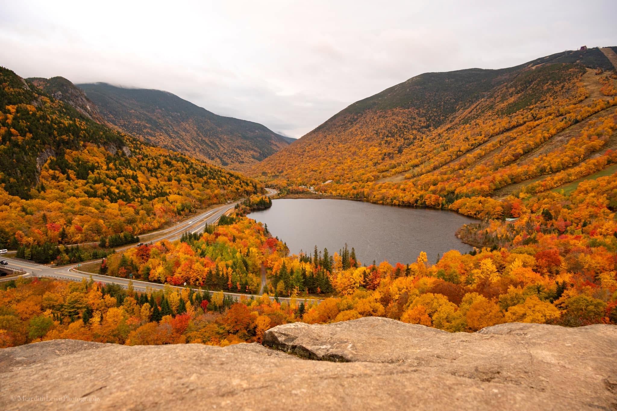 An elevated view of a New Hampshire notch in the White Mountains, with peak autumn foliage covering the mountainsides above a lake and a winding road.