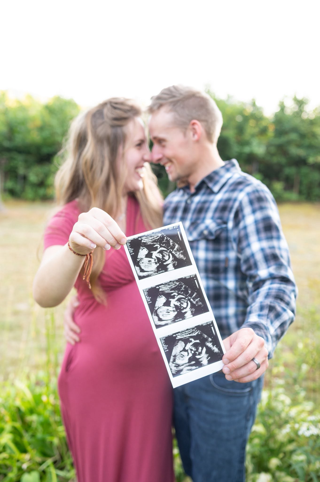 An expectant couple standing in a sunlit field, holding up a strip of black-and-white ultrasound photos.